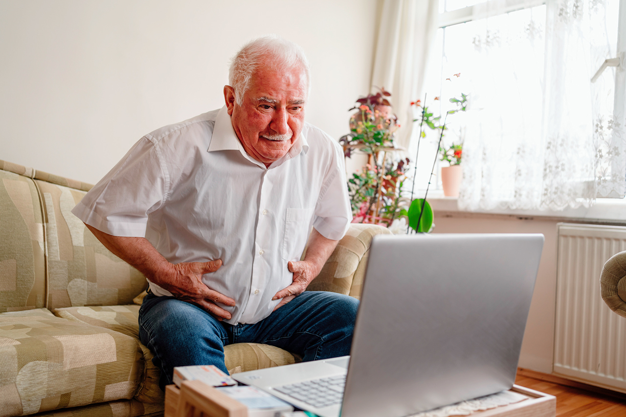 A senior patient looks distressed while holding his belly and looking at his computer.