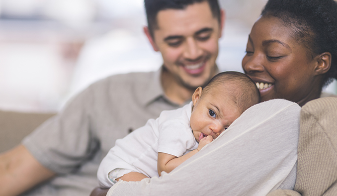 Una hermosa y joven madre afroamericana sujeta suavemente a su bebé contra su pecho. El bebé tiene la mano en la boca y los ojos bien abiertos. El papá está en el fondo con una gran sonrisa mientras se sienta en el sofá con su familia.