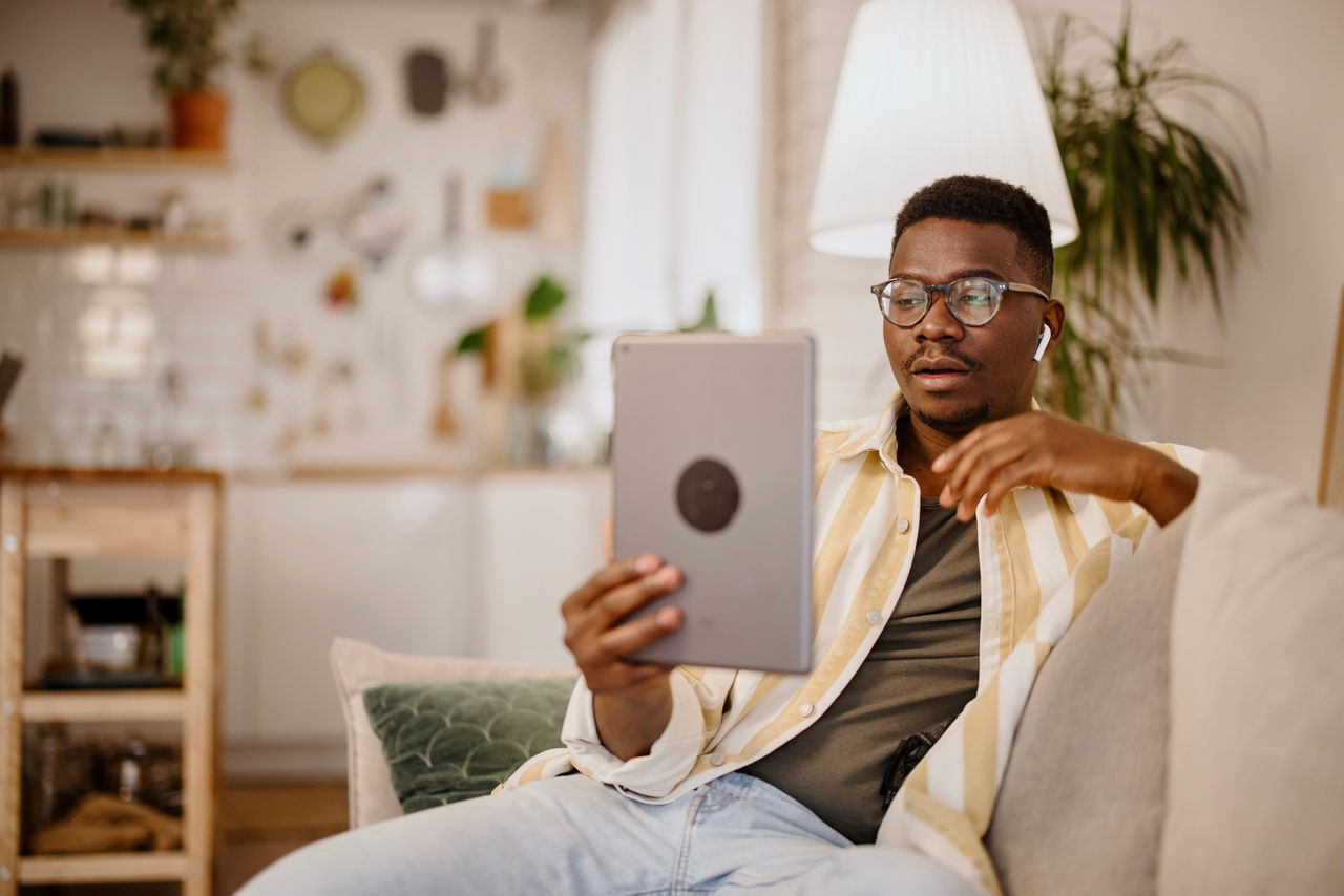 A Black man sites on a couch while reading from a tablet