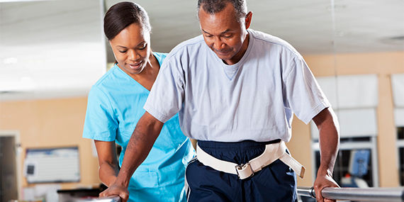 A man walks holding onto railings while a physical therapist looks on and assists