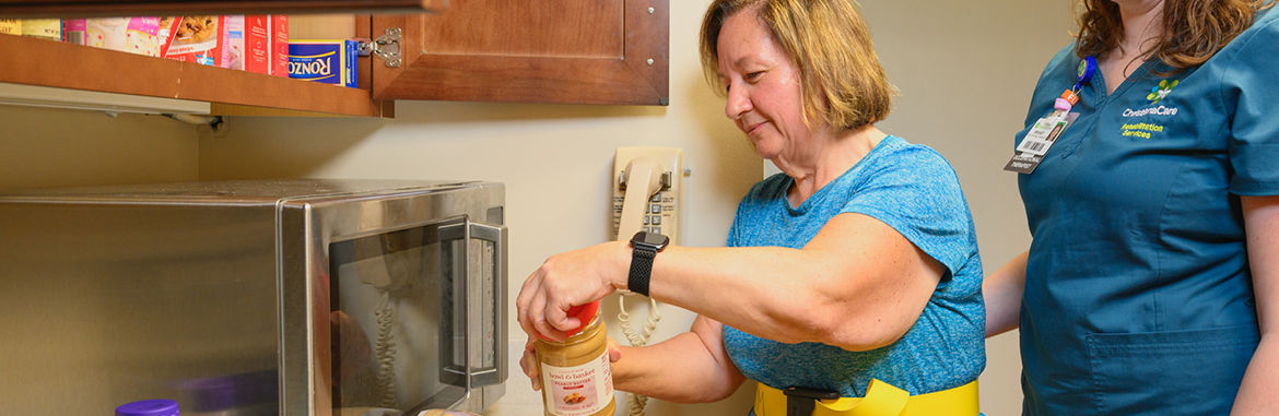 A female stroke patient stands in a kitchen opening a jar of peanut butter
