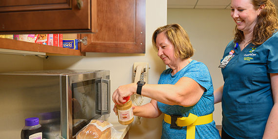 A female stroke patient stands in a kitchen opening a jar of peanut butter