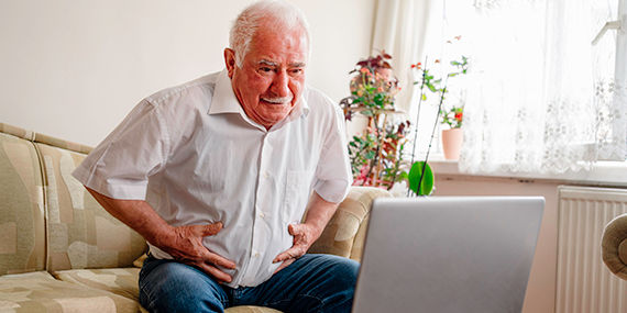 A senior patient looks distressed while holding his belly and looking at his computer.