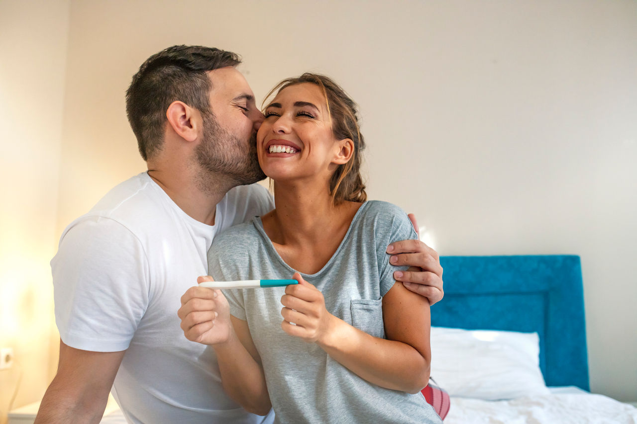 A woman smiles while holding a pregnancy test, while her partner hugs and kisses her.