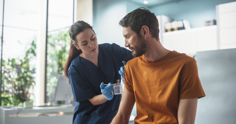 A man sits with his right sleeve rolled up watching while a female medical povider injects him with the flu vaccine