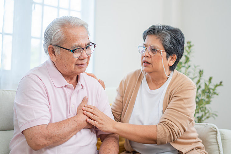 A senior Asian man holds his hand over his chest while his wife sits next to him, a look of concern on her face