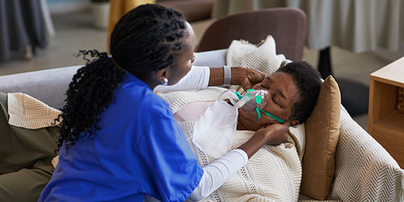 A nurse adjusts an oxygen mask on a patient's face. The patient is lying on her couch with her eyes closed.