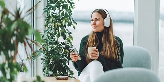A woman wearing headphones holds a glass and her phone while sitting in a chair.