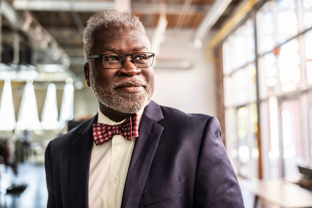 An older Black man wearing a suit jacket and bow tie smiles as he looks into the distance