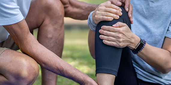 A woman sits around the ground holding her knee, while a man crouches next to her, comforting her