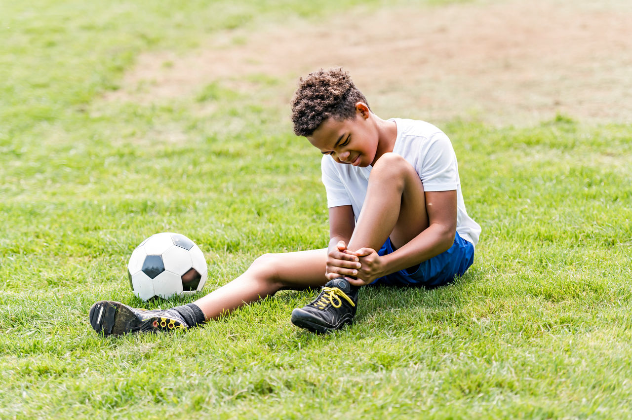 A child sits on the ground in pain holding his ankle with a soccer ball next to him.