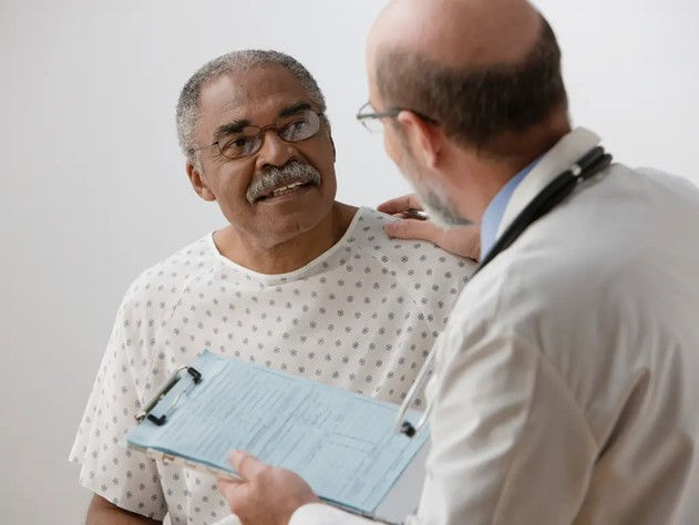 A Senior Black man wearing a hospital gown smiles at a doctor who is talking to him and touching his shoulder.
