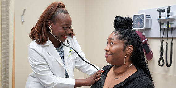 A Black female doctor uses a stethoscope on a young Black woman in an exam room.