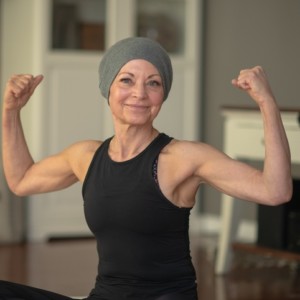 A senior woman with cancer flexes her muscles while sitting at home on her floor. She is smiling because she is getting stronger.