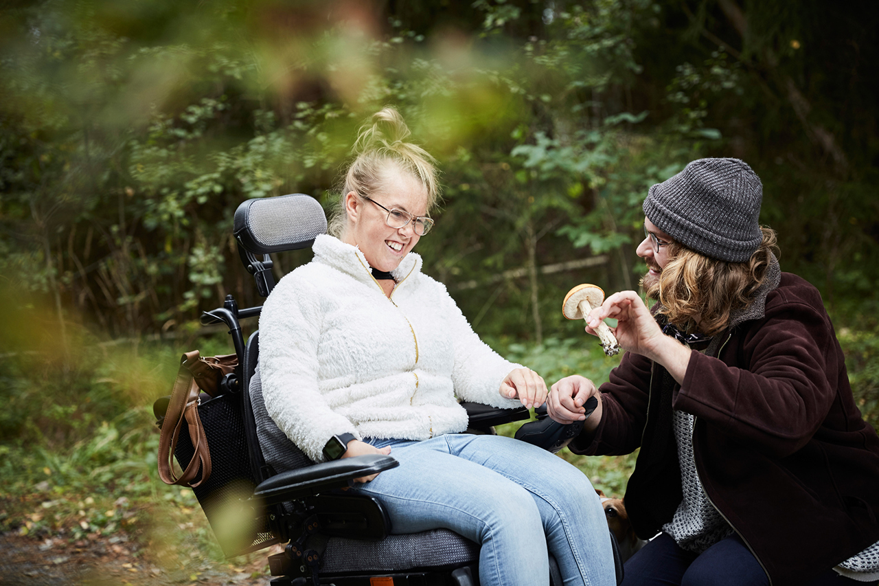 A male caretaker showing mushroom to a young woman with cerebral palsy in wheelchair in a forest.