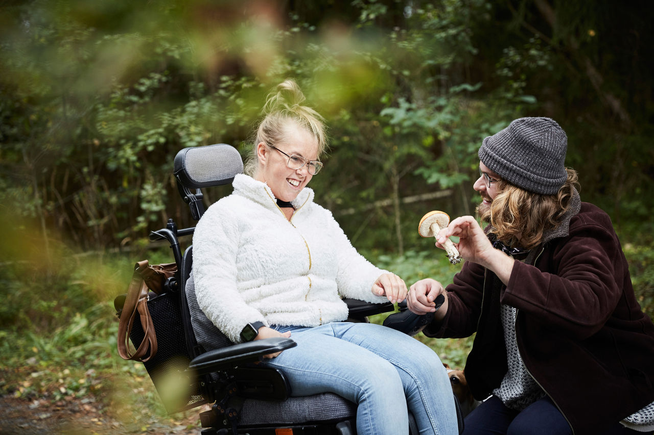 A male caretaker showing mushroom to a young woman with cerebral palsy in wheelchair in a forest.