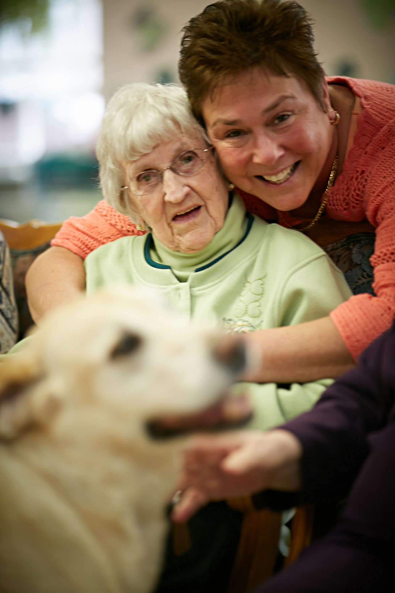 A caregiver hugs an elderly woman, with a dog in the foreground.
