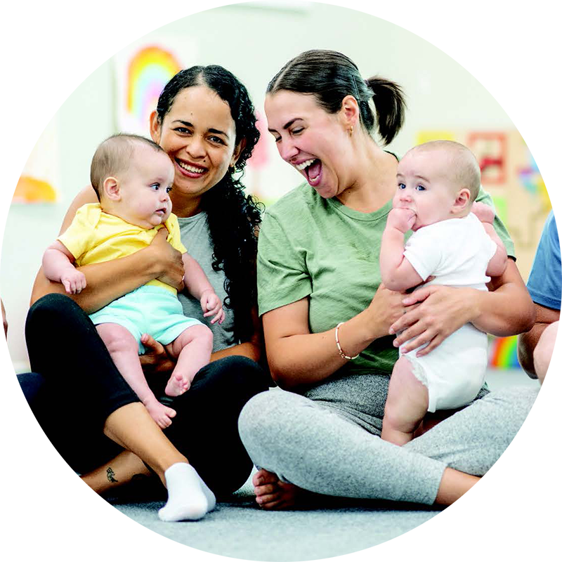 Two young mothers sit next to each other smiling and laughing while they hold their young babies