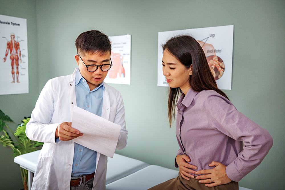 A young woman sits in an examination room holding her abdomen in distress while speaking to a medical professional