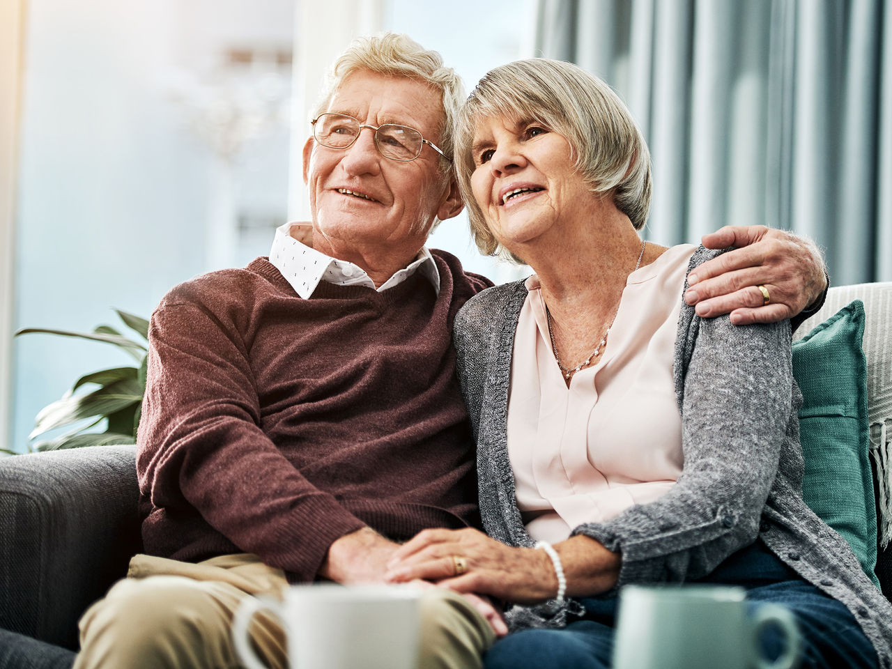 A senior couple sit holding each other while they look into the distance, with coffee mugs on the table in front of them