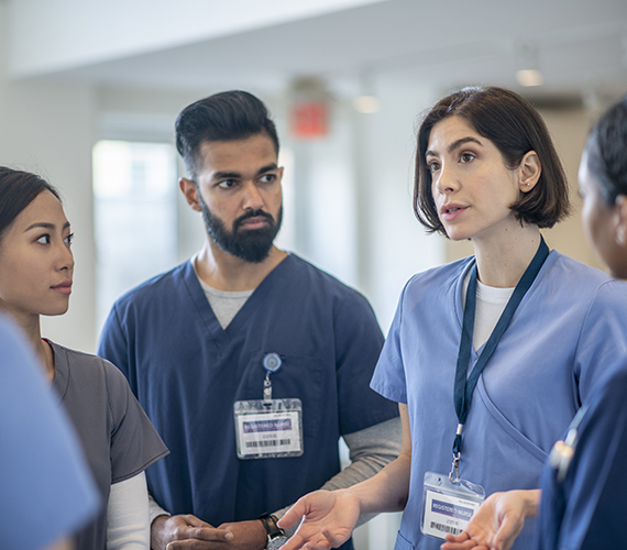 A small group of diverse medical professionals stand in the hallway for a brief meeting.  They are each wearing scrubs and focused on the conversation.