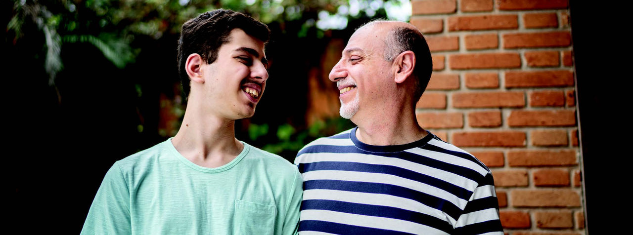A father and adult son stand shoulder to shoulder smiling at each other