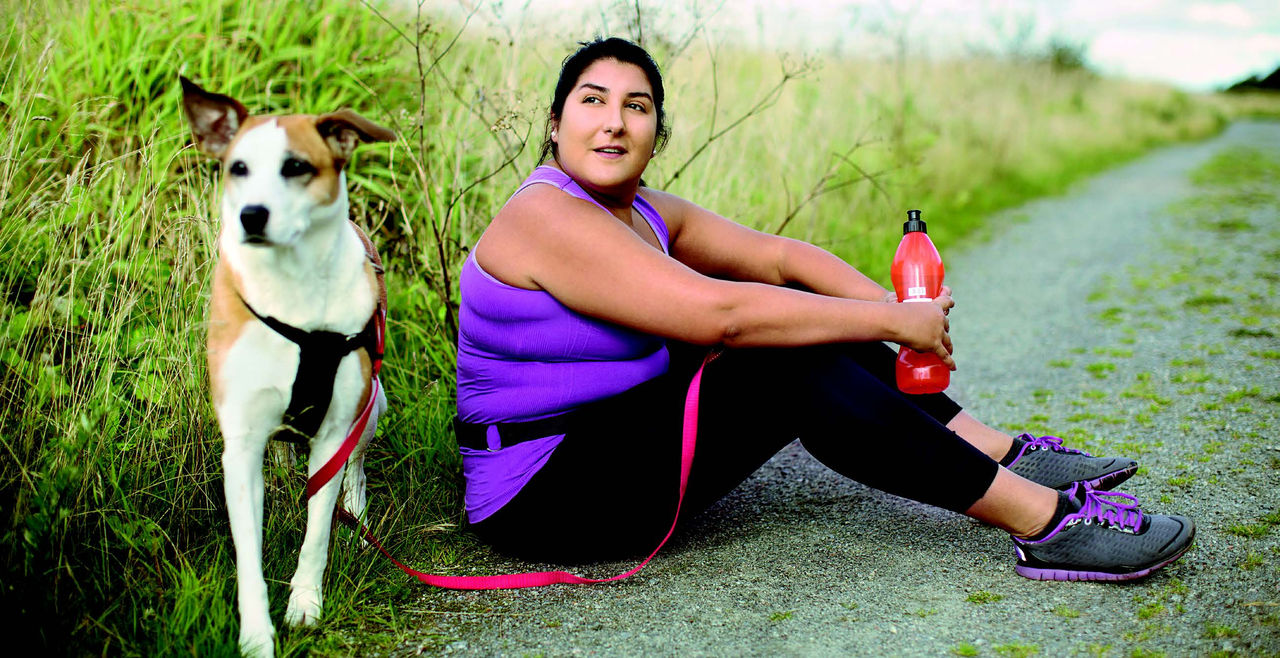 A woman in exercise-wear sites on a trail drinking from a water bottle, as her dog stands next to her