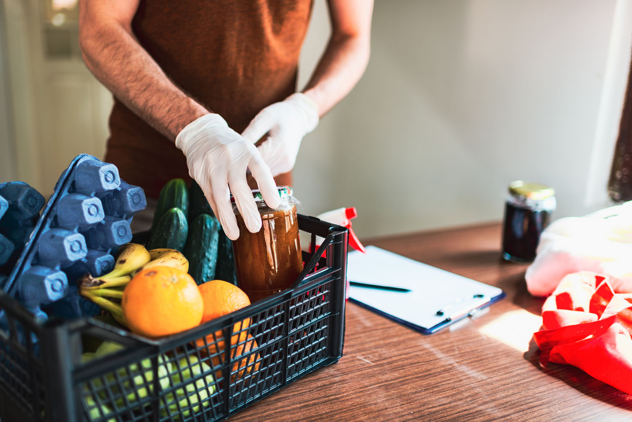 A man wearing rubber gloves collects groceries in a basket while checking a list