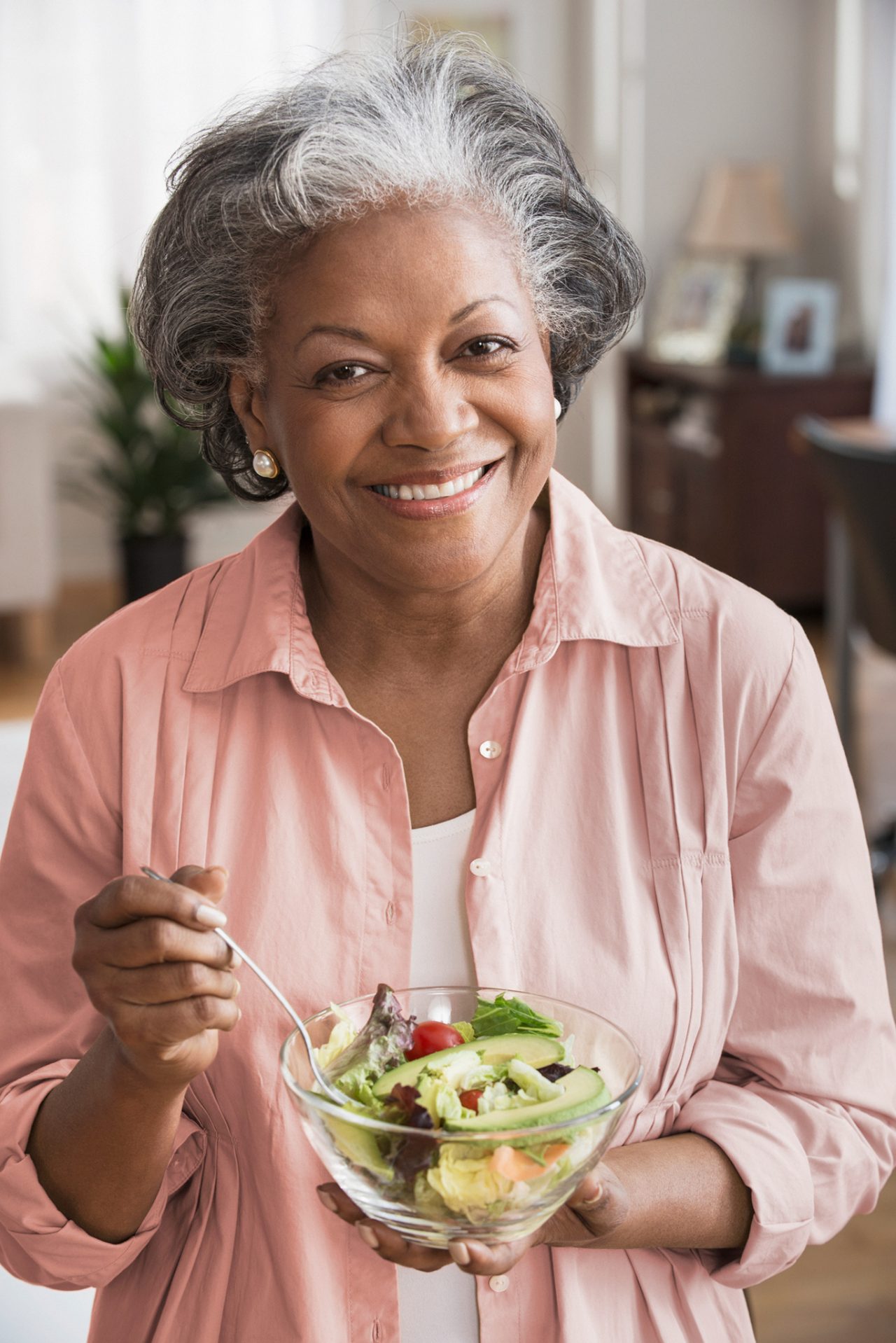 Una mujer negra de edad avanzada sonriente sostiene un tazón que contiene ensalada.